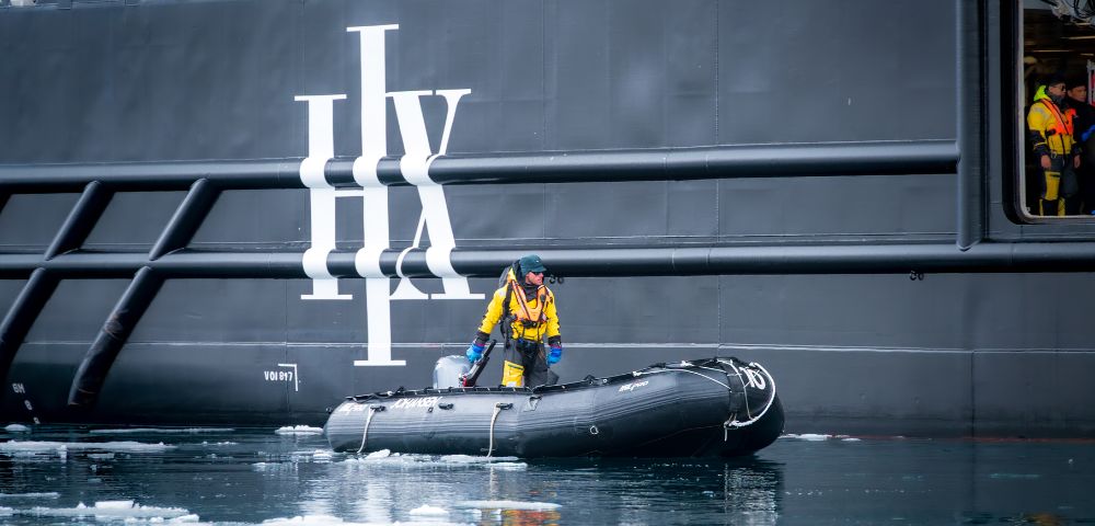 A person in a yellow waterproof suit navigates a small inflatable boat near a large dark vessel, surrounded by icy water.