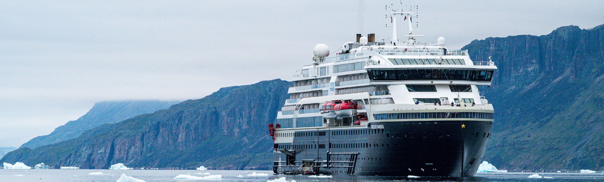 A large cruise ship sails through icy waters, flanked by towering green mountains under a cloudy sky. Icebergs float nearby.