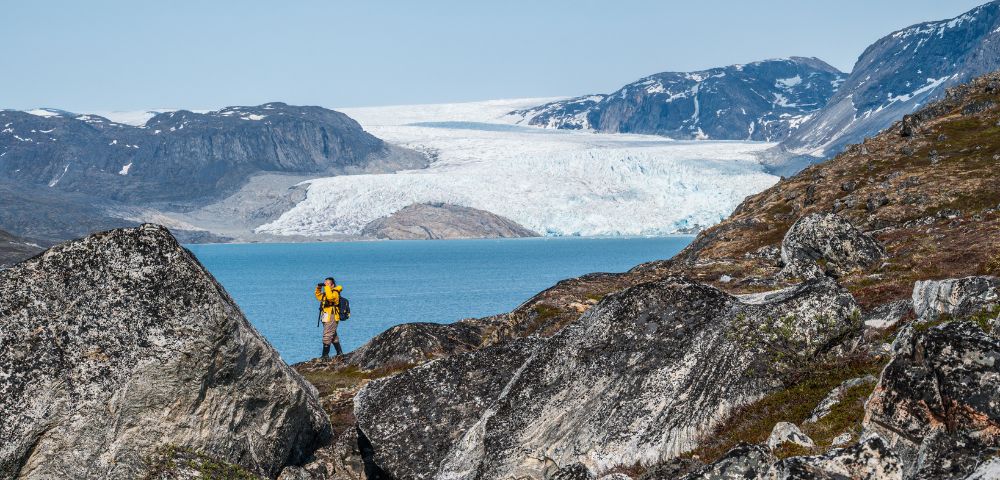 A hiker in a yellow jacket stands among boulders by a glacier-fed lake, with a vast glacier in the background under a clear blue sky.