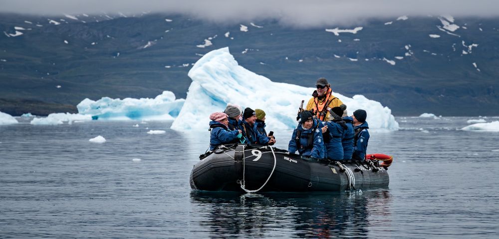 A group of people in winter gear aboard a black inflatable boat explore icy waters, with large icebergs in the background.