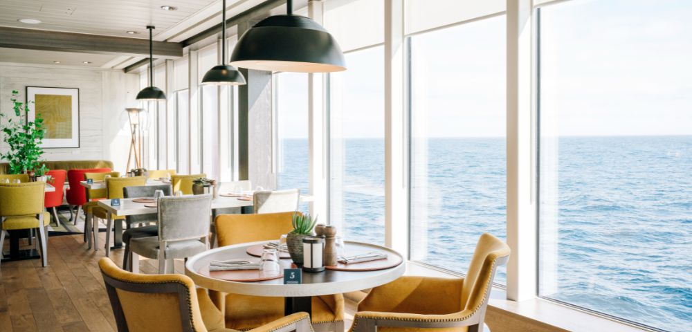Modern dining area with large windows overlooking the ocean, featuring neatly set tables, cushioned chairs, and pendant lighting.