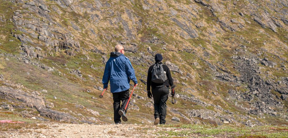 Two individuals walking along a winding mountain trail surrounded by greenery and rocky terrain.