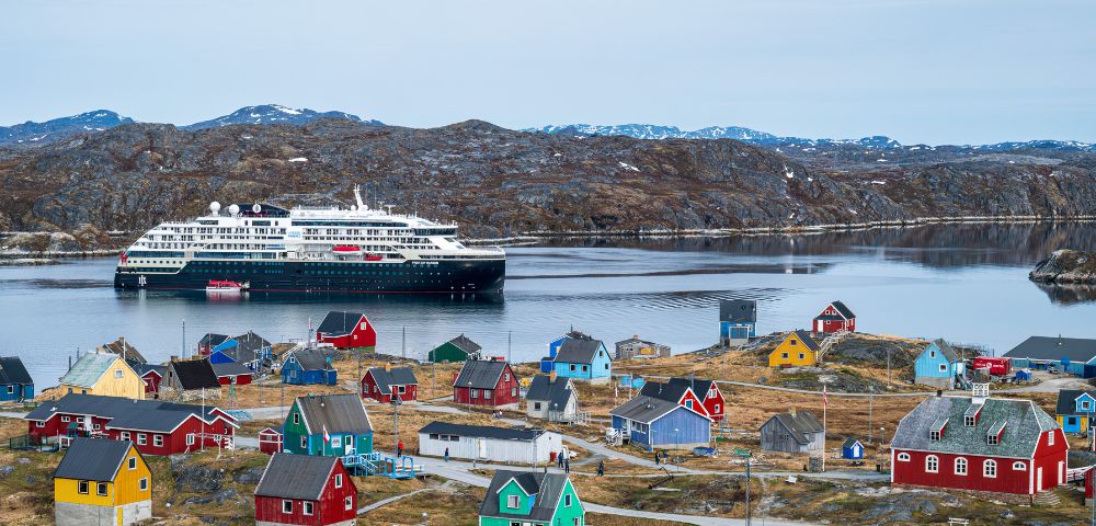 A cruise ship sails in the water near a quaint small town, surrounded by scenic landscapes and clear blue skies.