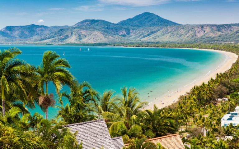 A stunning coastal view featuring a sandy beach, turquoise waters, lush palm trees, and distant mountains under a blue sky.