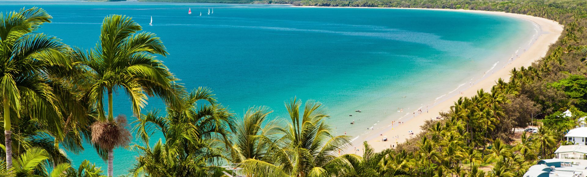 A stunning coastal view featuring a sandy beach, turquoise waters, lush palm trees, and distant mountains under a blue sky.