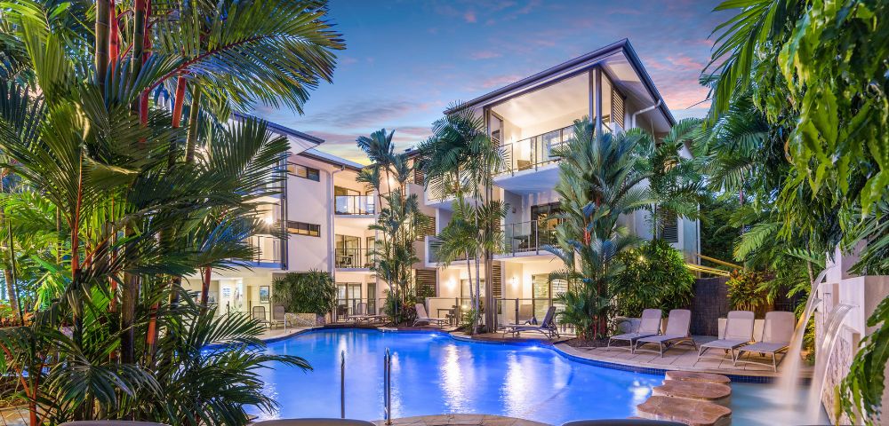 Sunset over the pool at a beachside resort, with vibrant colors reflecting on the water and palm trees in the background.
