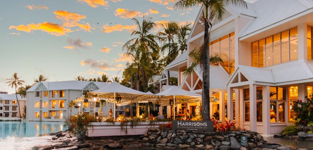 A beautiful sunset sky setting the tone for a warmly lit serene restaurant, surrounded by palm trees and on the edge of crystal clear water.