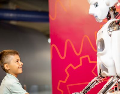 A young boy smiling and shaking hands with a humanoid robot against a vibrant pink background with orange patterns. The scene conveys curiosity and friendliness.