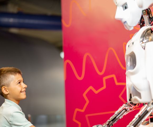 A young boy smiling and shaking hands with a humanoid robot against a vibrant pink background with orange patterns. The scene conveys curiosity and friendliness.