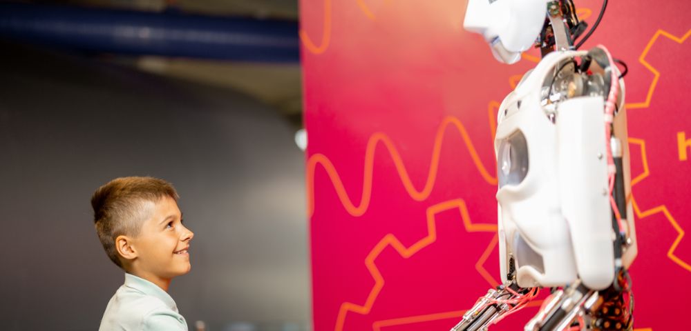 A young boy smiling and shaking hands with a humanoid robot against a vibrant pink background with orange patterns. The scene conveys curiosity and friendliness.
