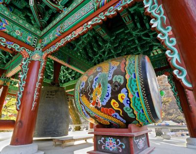 Vibrant Korean temple interior with intricate green, red, and blue patterns on wooden beams, featuring a large colorful drum and a bronze bell.