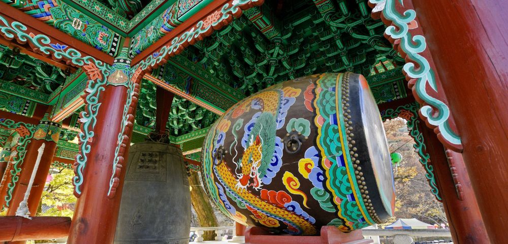 Vibrant Korean temple interior with intricate green, red, and blue patterns on wooden beams, featuring a large colorful drum and a bronze bell.