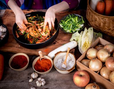 Hands mix ingredients for kimchi in a black bowl, surrounded by sliced green onions, cabbage, spices, garlic, onions, carrots, and an apple on a rustic table.