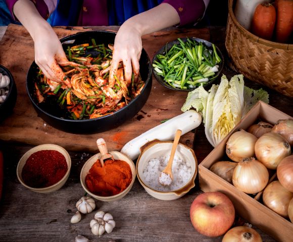 Hands mix ingredients for kimchi in a black bowl, surrounded by sliced green onions, cabbage, spices, garlic, onions, carrots, and an apple on a rustic table.