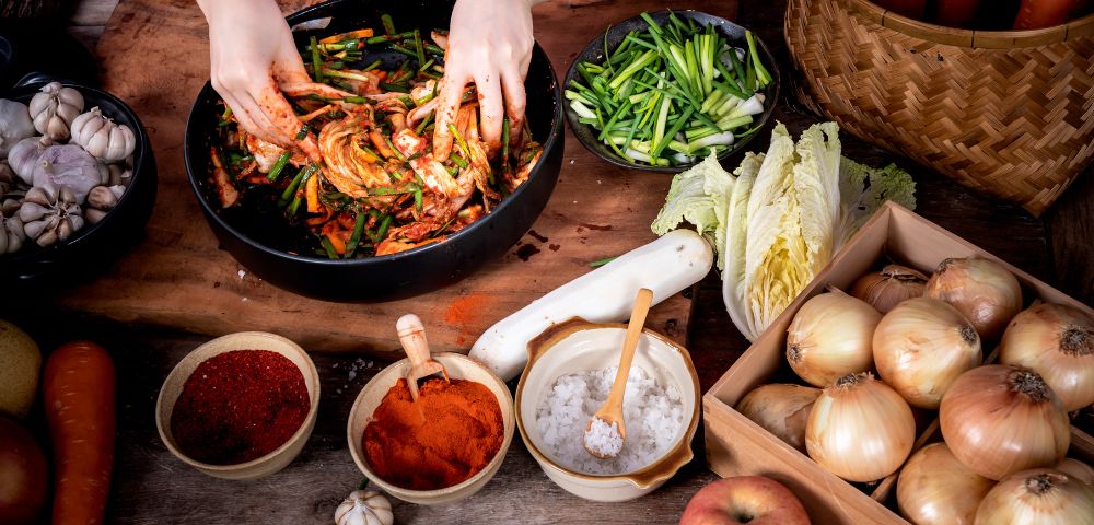 Hands mix ingredients for kimchi in a black bowl, surrounded by sliced green onions, cabbage, spices, garlic, onions, carrots, and an apple on a rustic table.
