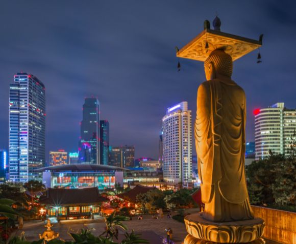 Golden Buddha statue overlooking illuminated modern cityscape at night. Skyscrapers light up the background, blending tradition and modernity.