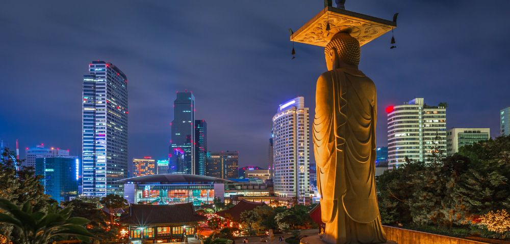 Golden Buddha statue overlooking illuminated modern cityscape at night. Skyscrapers light up the background, blending tradition and modernity.
