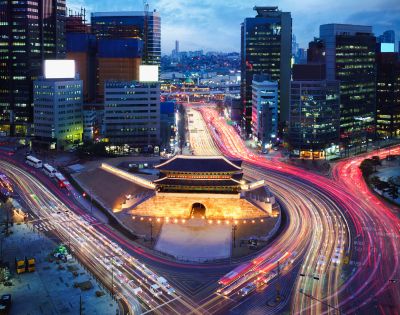 Aerial view of a vibrant cityscape at dusk, featuring light trails from traffic encircling an illuminated traditional gate, surrounded by modern skyscrapers.