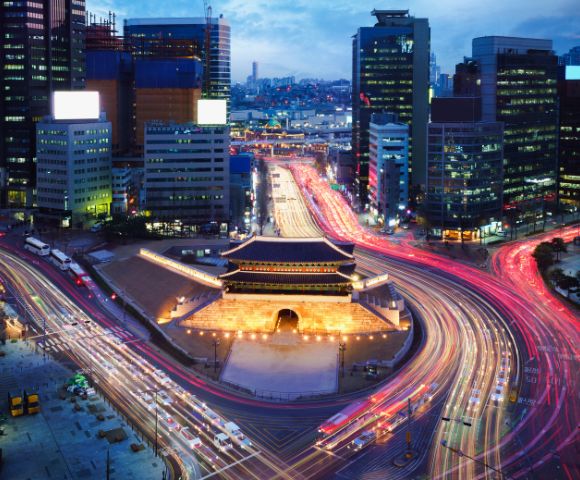 Aerial view of a vibrant cityscape at dusk, featuring light trails from traffic encircling an illuminated traditional gate, surrounded by modern skyscrapers.