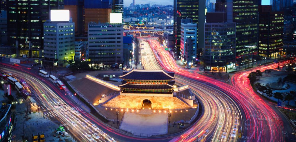 Aerial view of a vibrant cityscape at dusk, featuring light trails from traffic encircling an illuminated traditional gate, surrounded by modern skyscrapers.