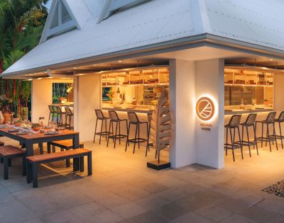 A spacious wooden table is set up outside, surrounded by benches and elegant tableware. The exterior features a sleek design with a brightly lit bar area, showcasing a welcoming vibe.