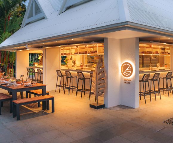 A spacious wooden table is set up outside, surrounded by benches and elegant tableware. The exterior features a sleek design with a brightly lit bar area, showcasing a welcoming vibe.