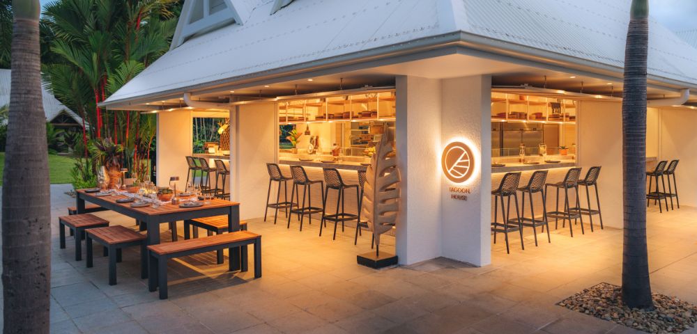A spacious wooden table is set up outside, surrounded by benches and elegant tableware. The exterior features a sleek design with a brightly lit bar area, showcasing a welcoming vibe.