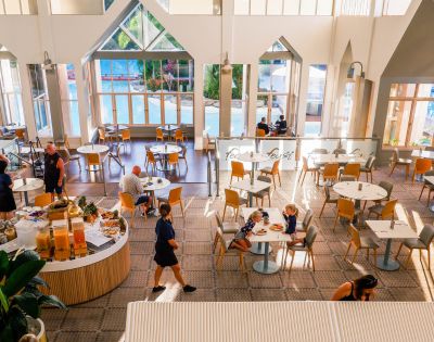 Bright, spacious dining area with high ceilings and large windows. Buffet tables with fruits, and people casually dining, creating a relaxed atmosphere.