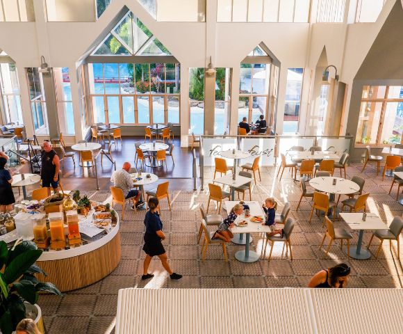 Bright, spacious dining area with high ceilings and large windows. Buffet tables with fruits, and people casually dining, creating a relaxed atmosphere.