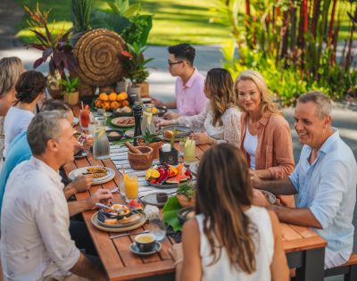 A group of people enjoys an outdoor meal at a long wooden table, surrounded by lush greenery. The scene is vibrant and cheerful. Plates of colorful fruit and drinks fill the table.