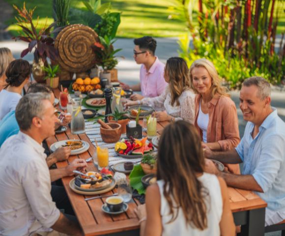 A group of people enjoys an outdoor meal at a long wooden table, surrounded by lush greenery. The scene is vibrant and cheerful. Plates of colorful fruit and drinks fill the table.