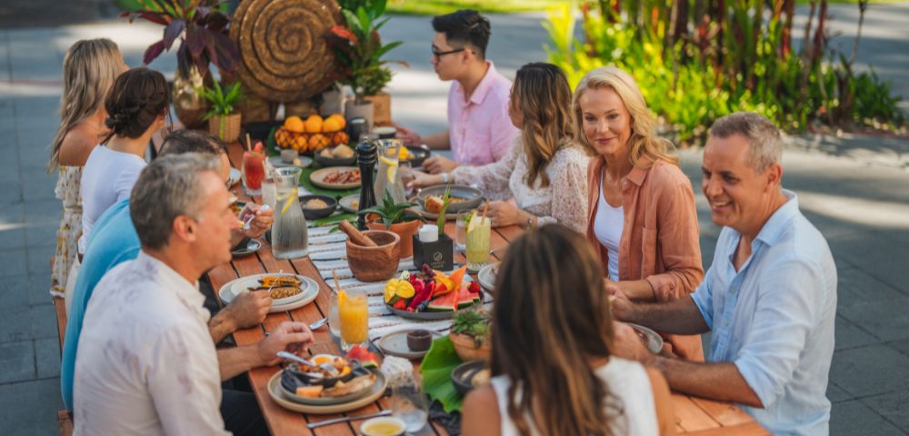 A group of people enjoys an outdoor meal at a long wooden table, surrounded by lush greenery. The scene is vibrant and cheerful. Plates of colorful fruit and drinks fill the table.
