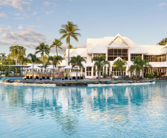 Luxurious tropical resort with a large, inviting pool surrounded by palm trees. Elegant white buildings and blue skies create a serene atmosphere.