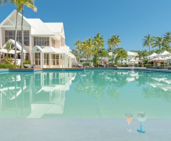 Luxurious tropical resort scene with a pristine pool, elegant white buildings, and lush palm trees under a clear blue sky. Two cocktails sit poolside.