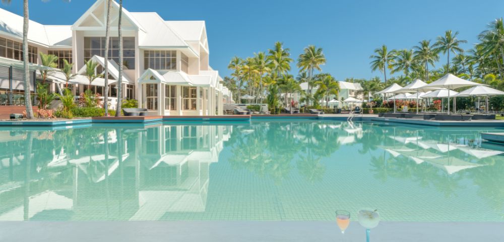Luxurious tropical resort scene with a pristine pool, elegant white buildings, and lush palm trees under a clear blue sky. Two cocktails sit poolside.