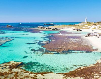 Clear turquoise waters with patches of seaweed lead to a sandy shoreline. A white lighthouse stands on a grassy hill under a bright blue sky.