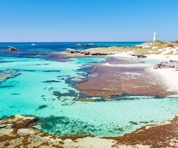 Clear turquoise waters with patches of seaweed lead to a sandy shoreline. A white lighthouse stands on a grassy hill under a bright blue sky.