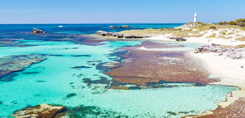 Clear turquoise waters with patches of seaweed lead to a sandy shoreline. A white lighthouse stands on a grassy hill under a bright blue sky.