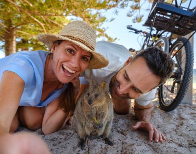 A couple, smiling and joyful, takes a selfie with a curious quokka on a sunny beach. Bicycles are in the background, and trees frame the scene.