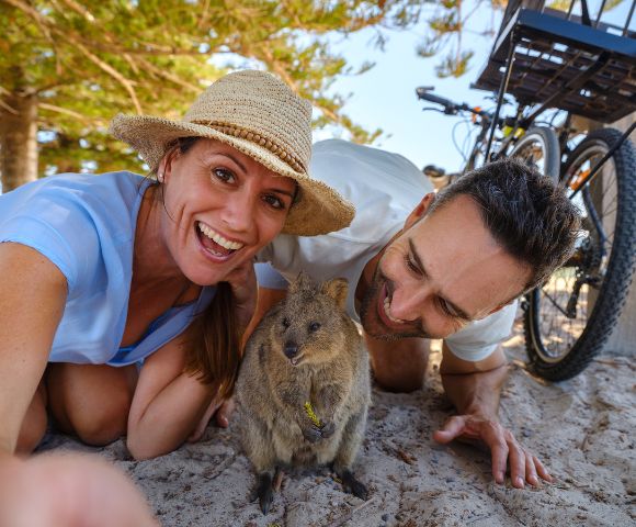 A couple, smiling and joyful, takes a selfie with a curious quokka on a sunny beach. Bicycles are in the background, and trees frame the scene.