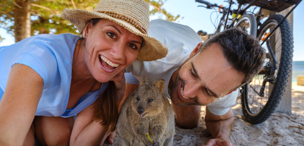 A couple, smiling and joyful, takes a selfie with a curious quokka on a sunny beach. Bicycles are in the background, and trees frame the scene.