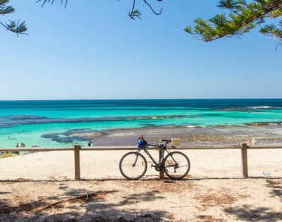 A tranquil beach scene with a bicycle leaned against a wooden fence in the foreground. Clear turquoise water and blue sky evoke a serene, sunny ambiance.