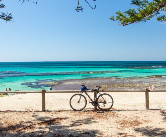 A tranquil beach scene with a bicycle leaned against a wooden fence in the foreground. Clear turquoise water and blue sky evoke a serene, sunny ambiance.