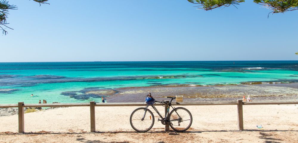 A tranquil beach scene with a bicycle leaned against a wooden fence in the foreground. Clear turquoise water and blue sky evoke a serene, sunny ambiance.
