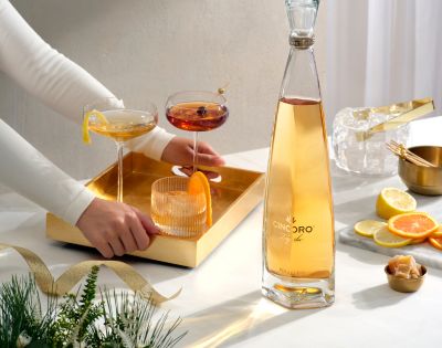 A person arranges a gold tray with cocktails beside a tall, sleek bottle on a table. Nearby are sliced citrus and festive greenery. Elegant and inviting scene.