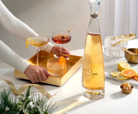A person arranges a gold tray with cocktails beside a tall, sleek bottle on a table. Nearby are sliced citrus and festive greenery. Elegant and inviting scene.