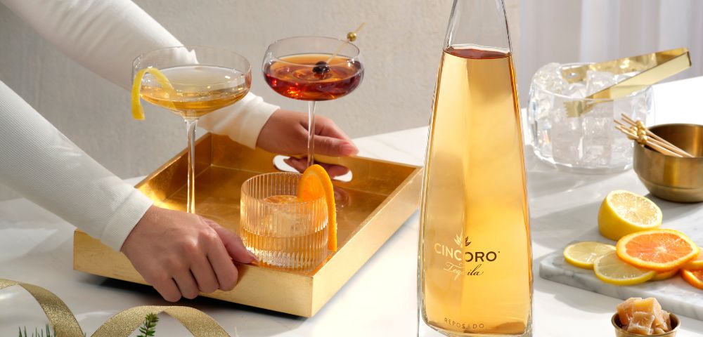 A person arranges a gold tray with cocktails beside a tall, sleek bottle on a table. Nearby are sliced citrus and festive greenery. Elegant and inviting scene.