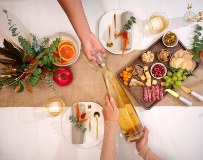 A festive table setting with two people sharing a large bottle of champagne. There's a charcuterie board, autumn decor, and elegant tableware.