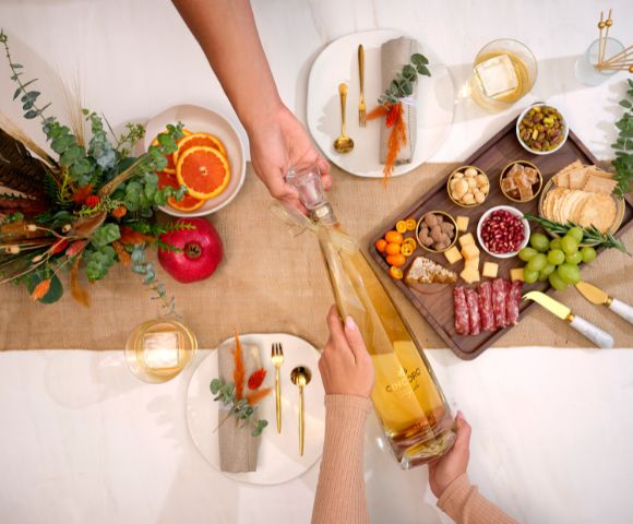 A festive table setting with two people sharing a large bottle of champagne. There's a charcuterie board, autumn decor, and elegant tableware.