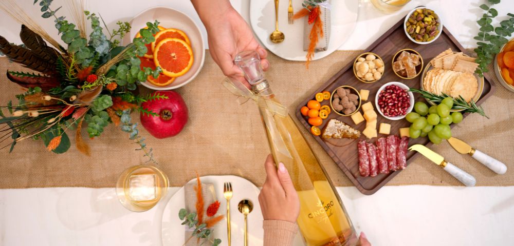 A festive table setting with two people sharing a large bottle of champagne. There's a charcuterie board, autumn decor, and elegant tableware.
